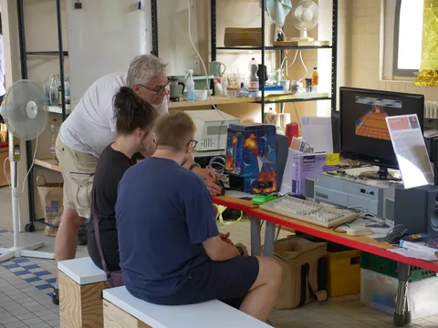 Drei Personen arbeiten zusammen an einem Computer in einem Werkstattraum mit Regalen und Technik.