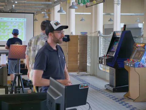 Mann mit Cap und Brille steht neben Laptop vor Arcade-Spielautomaten im Raum.