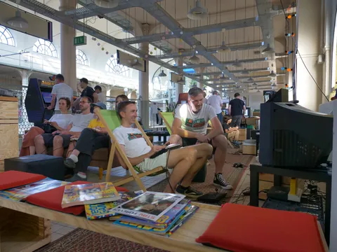 Gruppe spielt Videospiele vor einem alten Röhrenfernseher in einer hellen Halle mit hohen Decken.