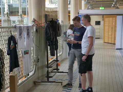 Zwei Männer betrachten ein Smartphone neben einem Stand mit T-Shirts in einer Halle.