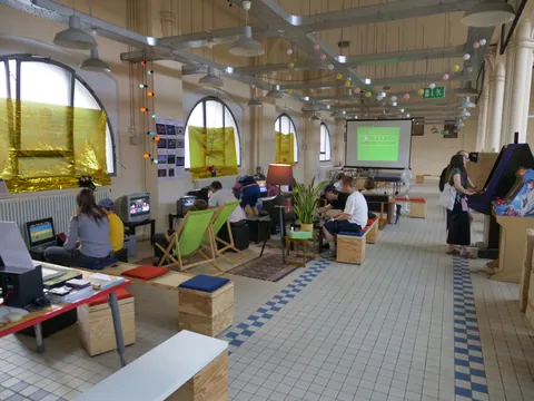 Menschen sitzen in einem großen, hellen Raum mit Computern, Arcade-Automaten und hoher Decke.
