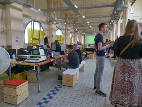 Menschen in einem hellen Atelier mit vintage Computern und hohen Fenstern.