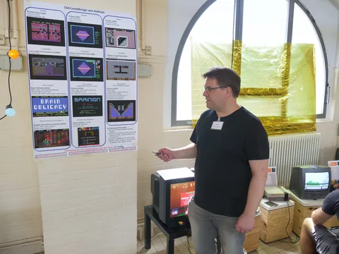 Mann präsentiert Poster mit C64-Levelentwürfen und Spielscreenshots neben Retro-Monitoren.