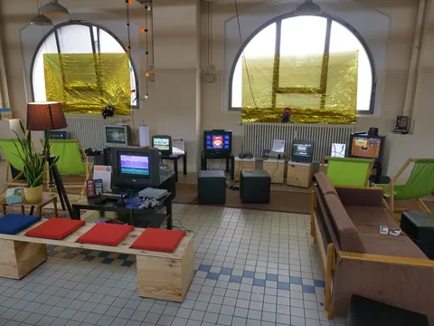 Retro-Gaming-Raum mit mehreren CRT-Monitoren, grünen Sesseln, Sofa, Spielkonsolen und großen Rundbogenfenstern.