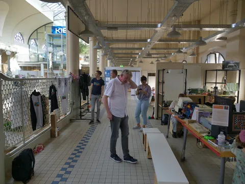 Innenansicht einer Markthalle mit Besuchern, Textilständen und Computertischen.