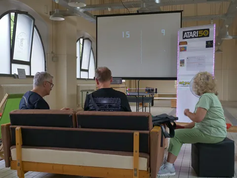 Drei Personen beobachten eine Pong-Projektion auf Leinwand neben einem Atari-50-Ausstellungsbanner.