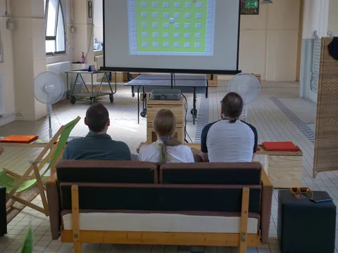 Drei Personen sitzen auf einem Sofa vor einer Projektionsleinwand mit grünem Rastermuster.