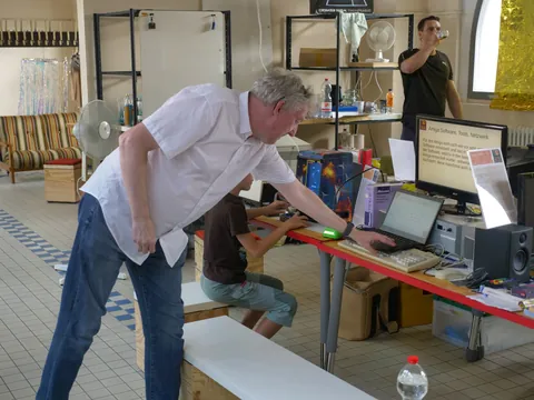 Mann im weißen Hemd bedient Laptop am Tisch, Kind sitzt dabei in einer Werkstatt.