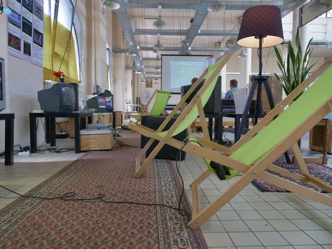 Grüne Deckchairs im Vordergrund eines Büros mit Retro-TV-Geräten, Schreibtischen und Stehlampe.