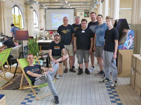 Acht Männer in einem kreativen Arbeitsraum mit Retro-Computern, Beamer und Arcade-Automaten.