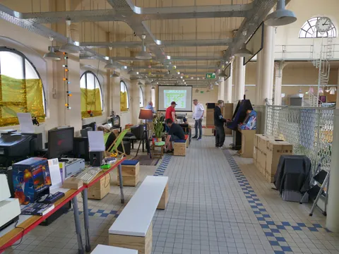 Hohe Industriehalle mit Retro-Computern, Arcade-Automaten, Beamer-Projektion und Besuchern.