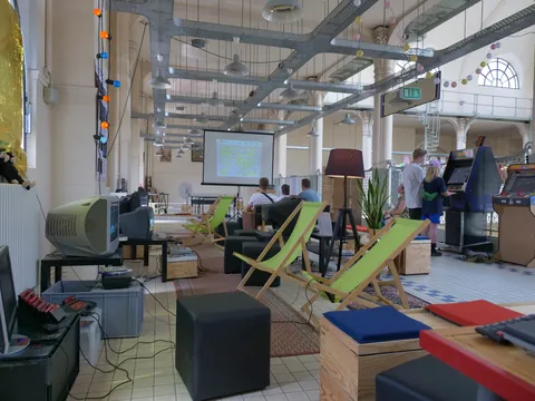 Retro-Gaming-Lounge mit alten CRT-Monitoren, grünen Liegestühlen, Arcade-Automaten und großer Projektionsleinwand.