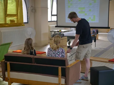 Mann zeigt auf eine Projektionswand während zwei Frauen auf einem Sofa zuschauen.