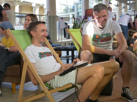 Zwei lächelnde Männer in weißen „Metro Computer Dresden“-Shirts sitzen in Liegestühlen und spielen ein Videospiel.