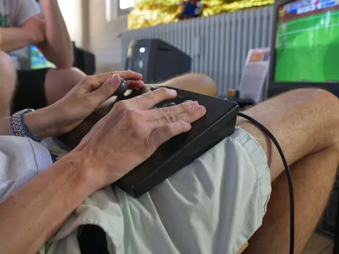 Hände bedienen einen schwarzen Joystick mit Trackball beim Videospielen vor einem Monitor.