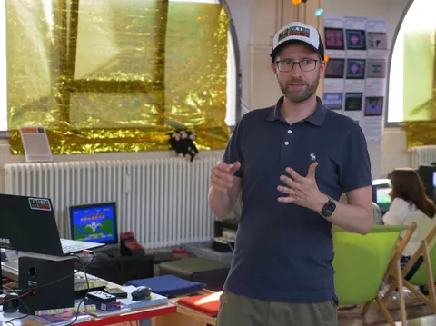 Mann mit Cap und Brille gestikuliert vor Laptop und Monitor im Arbeitsraum.