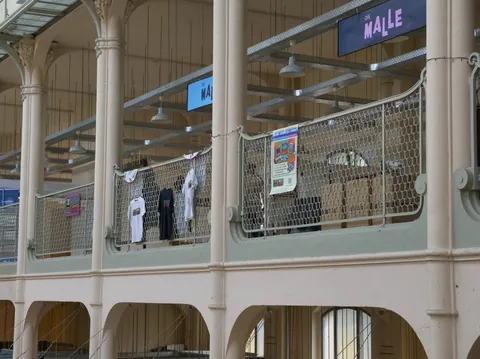 Innenraum mit weißen Säulen, Galerie mit Metallgeländer, hängenden T-Shirts und Schild „DIE MALLE“.