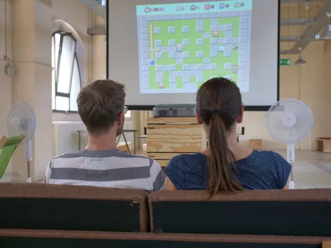 Zwei Personen auf einem Sofa betrachten eine Videospiel-Projektion an der Wand.