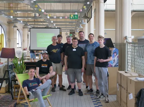 Gruppenfoto von acht Männern in einem hellen Veranstaltungsraum mit Projektionswand im Hintergrund.