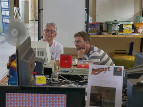 Zwei Männer sitzen vor einem Schreibtisch voller alter Computer, Kabeln und technischem Equipment.