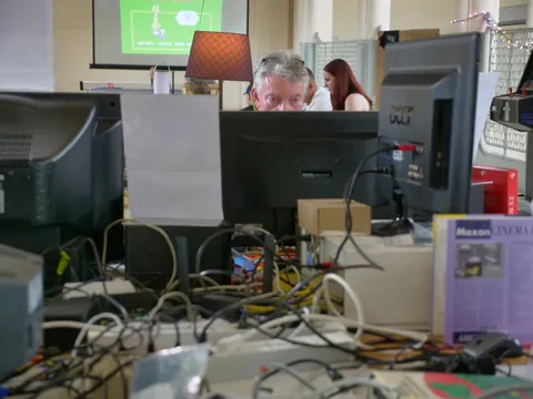 Mann blickt über einen Monitor in einem unordentlichen Büro mit Computern und Kabeln.