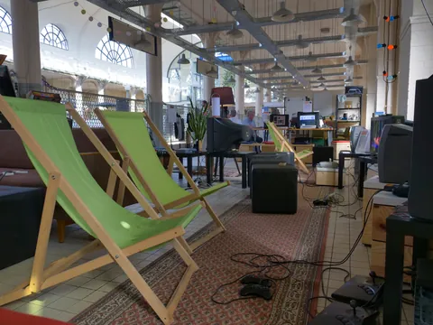 Grüne Liegestühle, alte CRT-Monitore und Gamecontroller in einer hellen Industriehalle mit Bogenfenstern.