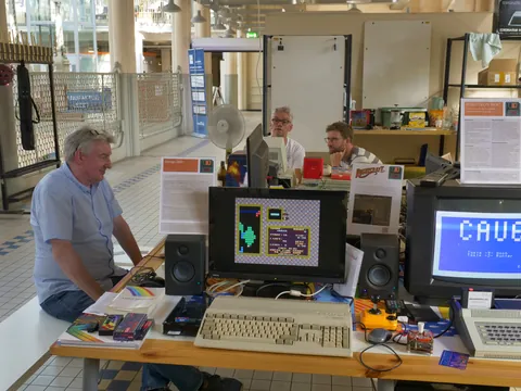 Drei Männer an einem Tisch mit Vintage-Computern, Monitoren und Retro-Videospielen.