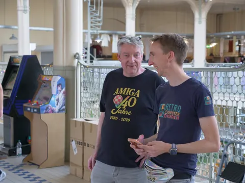 Zwei Männer in Amiga- und Retro-Computer-T-Shirts betrachten ein Smartphone neben Arcade-Automaten.