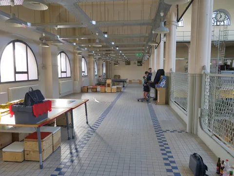 Geräumige Industriehalle mit hohen Decken, Bogenfenstern, kariertem Fliesenboden, Tischen und zwei Personen.