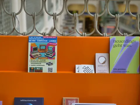 Orangefarbenes Regal mit Flyern zur Retro Computer Lounge und weiteren Veranstaltungskarten.