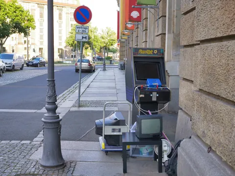 Arcade-Spielautomat und alte Fernsehgeräte stehen auf einem städtischen Gehweg neben einem Parkverbotsschild.