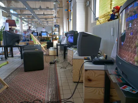 Vintage-Spielecke mit zahlreichen Röhrenfernsehern, Retro-Konsolen und Mario-Plüschtier in einem industriellen Raum.