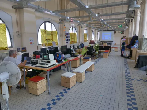 Heller Ausstellungsraum mit Retro-Computern, Arcade-Automaten und Besuchern vor einer Leinwand in historischem Gebäude.