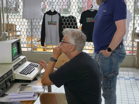 Mann mit Brille an altem Commodore-Computer, stehender Mann in blauem Shirt beobachtet.