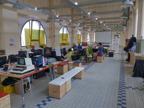 Heller, offener Arbeitsraum mit Computern, Retro-Geräten, Holzkisten und arbeitenden Personen vor hohen Rundbogenfenstern.