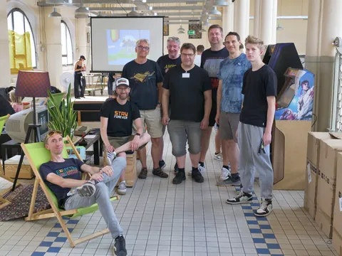 Acht Männer posieren in einem hellen Saal mit Projektor und Arcade-Automaten im Hintergrund.