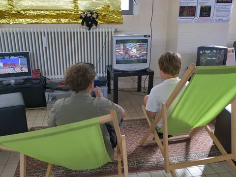 Zwei Jungen in grünen Liegestühlen spielen Videospiele auf einem Röhrenfernseher und einem Monitor.