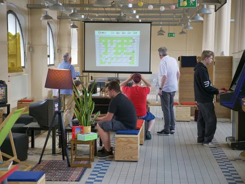 Mehrere Personen vor einer Projektionswand mit Tabellenkalkulation in einem Raum mit Retro-Spielgeräten.