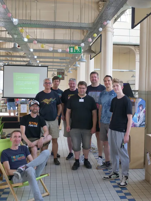 Gruppe Männer posiert in einem hellen Saal mit Lichterketten und Projektionsfläche.