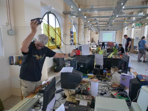 Grauhaariger Mann hält elektronisches Gerät über einem chaotischen Tisch voller alter Computer und Kabel.