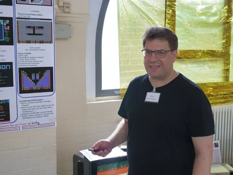 Mann mit Brille und Namensschild vor Poster zum C64-Spiel Pieces Level Editor.