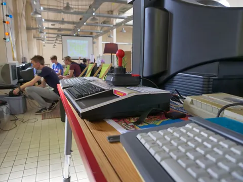 Retrocomputer mit rotem Joystick im Vordergrund, im Hintergrund Menschen bei einem Retro-Gaming-Treffen.