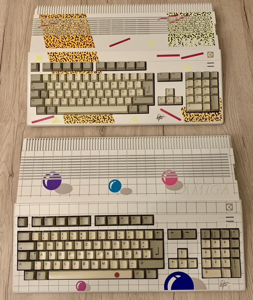 Draufsicht auf zwei originale Amiga 500 der „New Art Edition", die auf einem hellen Holzboden liegen. Das obere Gerät zeigt das „Leopard-Design" mit gelb-schwarzen Mustern und pinken Streifen. Das untere Gerät zeigt das „Ball-Design" mit bunten Kugeln auf einem grauen Gitter-Hintergrund. Beide Gehäuse tragen unten rechts die aufgedruckte Unterschrift von Stefanie Tücking.